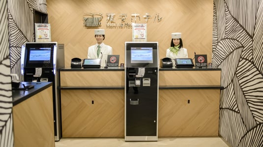 Two humanoid robots in white uniforms and hats stand behind a hotel check-in desk.