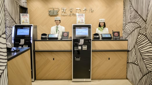 Two humanoid robots in white uniforms and hats stand behind a hotel check-in desk.
