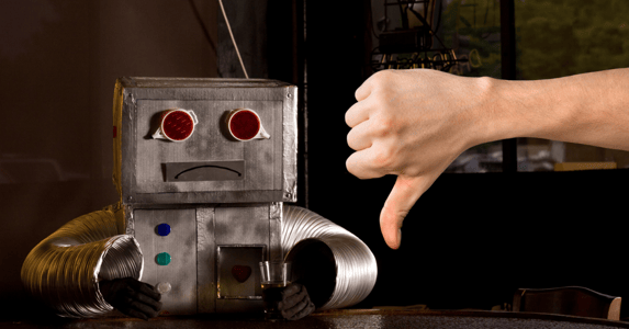 A robot with a frown sits at a table in front of a person's hand making a thumbs down sign.