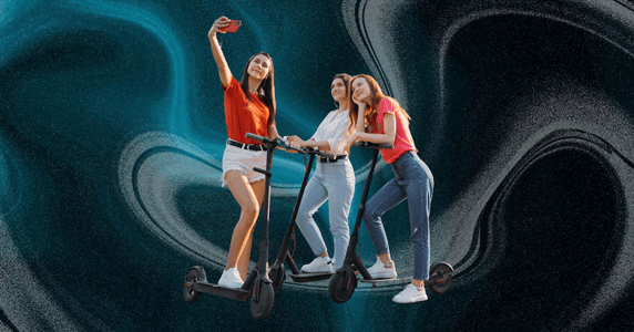 Three women take a selfie while riding e-scooters.