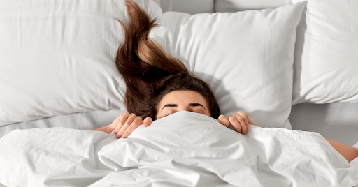 A person with long hair pulling a blanket over their head while laying in bed.