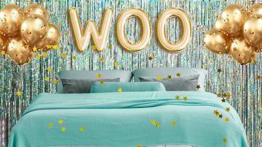 A bed topped with a teal bedspread and gray pillows surrounded by gold balloons, including three gold balloons that spell out, 