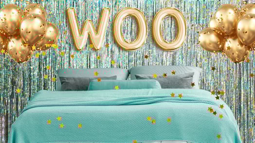A bed topped with a teal bedspread and gray pillows surrounded by gold balloons, including three gold balloons that spell out, 
